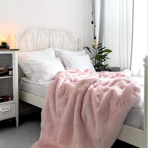 May include: A pink faux fur throw blanket draped over a white bed with a white metal headboard. The blanket is soft and fluffy, and it would be perfect for adding a touch of luxury to any bedroom.