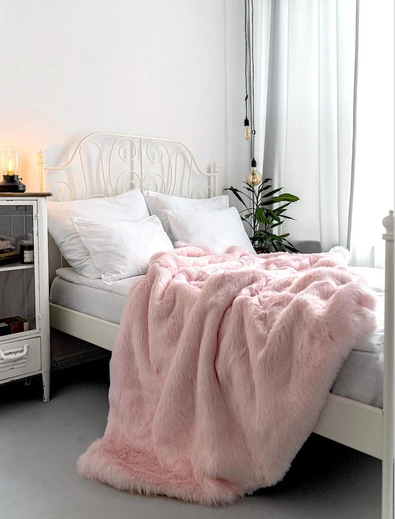 May include: A pink faux fur throw blanket draped over a white bed with a white metal headboard. The blanket is soft and fluffy, and it would be perfect for adding a touch of luxury to any bedroom.