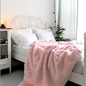 Pode incluir: Uma cama com uma cabeceira de metal branco e uma manta de pele sintética rosa. A cama é feita com roupa de cama e almofadas brancas.