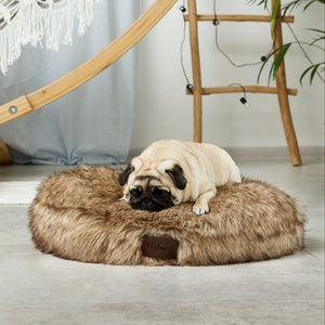 May include: A brown faux fur dog bed with a round shape. A light brown pug dog is lying on the bed. The bed has a brown leather tag with the word "Wilson" on it.