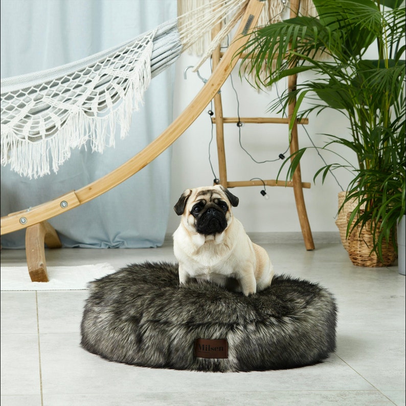 May include: A gray and black faux fur dog bed with a brown leather label that says "Aillsen". A pug dog is sitting on the bed.