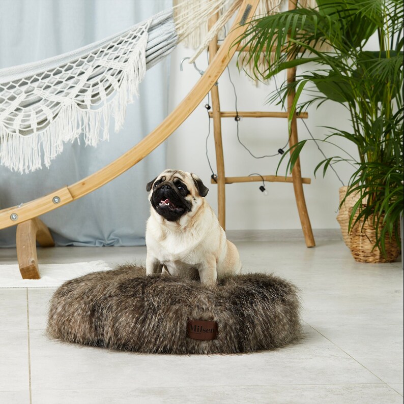 May include: A brown and gray faux fur dog bed with a brown leather tag that says "Milsa". A pug dog is sitting on the bed.