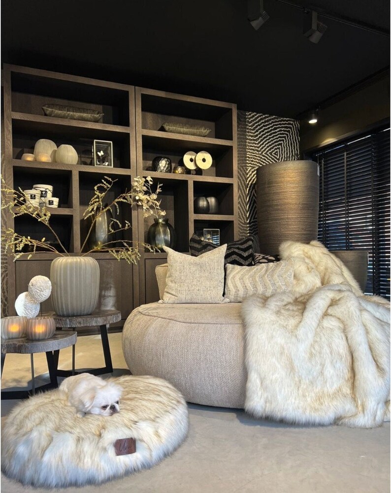 May include: A light brown, round, faux fur dog bed with a white dog lying on it. The bed is in a living room with a large, round, light brown sofa with a white faux fur throw blanket on it. There are two dark brown wooden shelves with various decorative items on them.