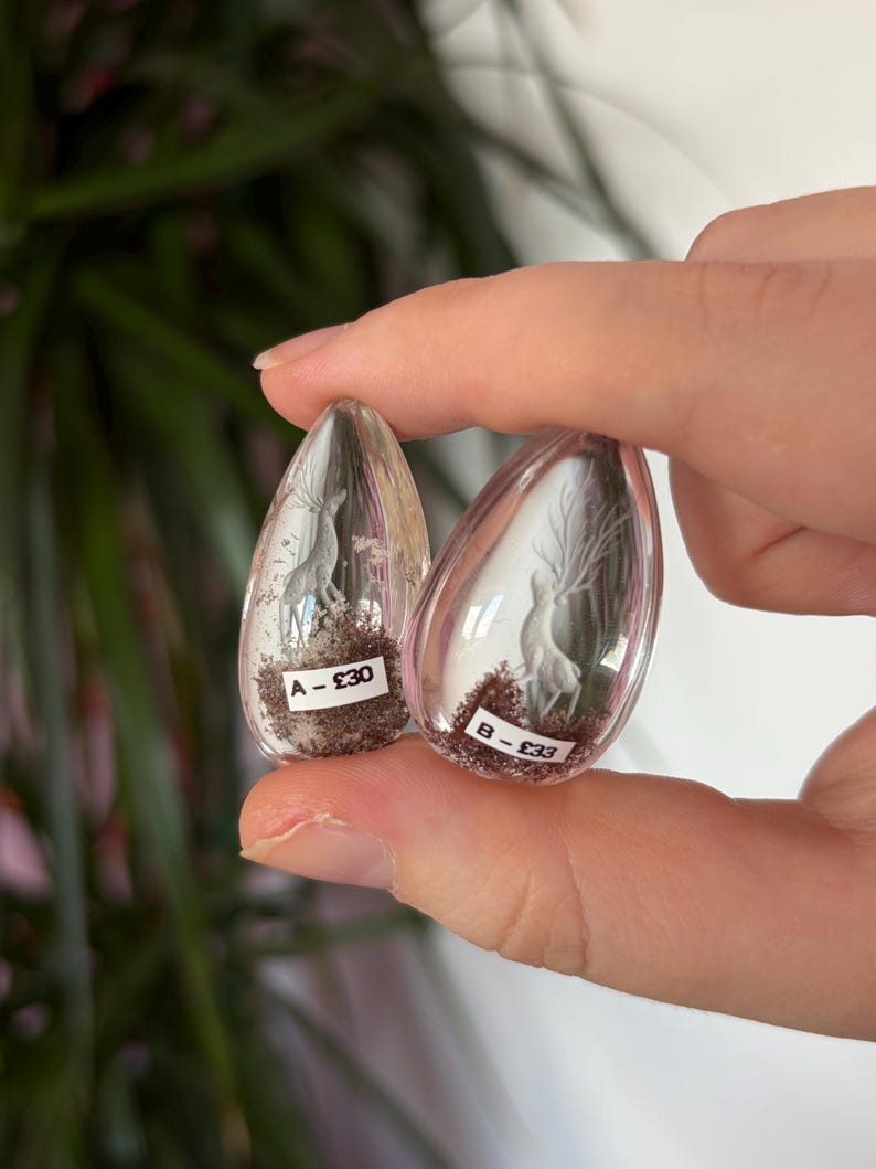 K&ouml;nnte beinhalten: Zwei transparente, tropfenf&ouml;rmige Harzornamente mit Miniatur-Hirschfiguren und dunkelbraunem, strukturiertem Material an der Basis. Jedes St&uuml;ck hat ein kleines wei&szlig;es Etikett mit schwarzem Text, eines mit A - &pound;30 und das andere B - &pound;33.