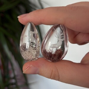 K&ouml;nnte beinhalten: Zwei transparente, tropfenf&ouml;rmige Harzanh&auml;nger mit eingebetteten wei&szlig;en Hirschfiguren. Der linke Anh&auml;nger enth&auml;lt hellgraues, strukturiertes Material an der Basis, w&auml;hrend der rechte Anh&auml;nger ein dunkelrotbraunes, moosartiges Basismaterial aufweist.