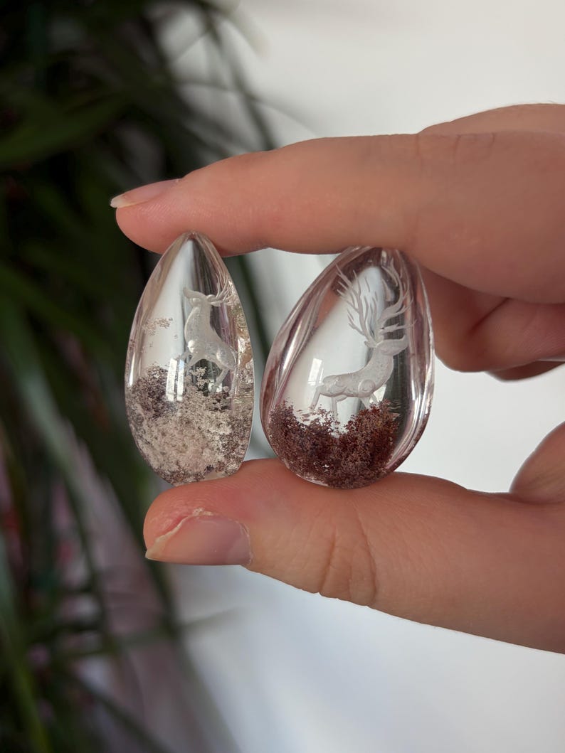 K&ouml;nnte beinhalten: Zwei transparente, tropfenf&ouml;rmige Harzanh&auml;nger mit eingebetteten wei&szlig;en Hirschfiguren. Der linke Anh&auml;nger enth&auml;lt hellgraues, strukturiertes Material an der Basis, w&auml;hrend der rechte Anh&auml;nger ein dunkelrotbraunes, moosartiges Basismaterial aufweist.