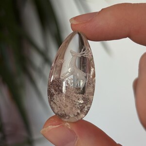 K&ouml;nnte beinhalten: Ein tropfenf&ouml;rmiger Anh&auml;nger aus klarem Quarz mit einer eingravierten Hirschfigur mit Geweih. Die Basis des Steins enth&auml;lt nat&uuml;rliche braune und wei&szlig;e Mineraleinschl&uuml;sse, die ein strukturiertes, erdiges Aussehen im transparenten Kristall erzeugen.