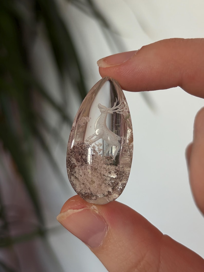 K&ouml;nnte beinhalten: Ein tropfenf&ouml;rmiger Anh&auml;nger aus klarem Quarz mit einer eingravierten Hirschfigur mit Geweih. Die Basis des Steins enth&auml;lt nat&uuml;rliche braune und wei&szlig;e Mineraleinschl&uuml;sse, die ein strukturiertes, erdiges Aussehen im transparenten Kristall erzeugen.