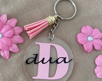 Op de afbeelding: Een roze acryl sleutelhanger met de letter "D" en het woord "dua" in het zwart. De sleutelhanger heeft een roze kwastje aan een zilveren ketting.