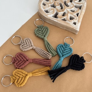 May include: A selection of macrame heart keyrings in shades of green, beige, red, yellow, blue, and black. Each keyring has a heart-shaped design with a tassel and a silver ring. A wooden heart decoration is in the background.
