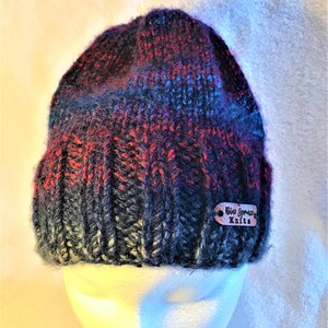 May include: A hand-knitted beanie hat in a mix of colors including red, blue, and black. The hat is displayed on a white mannequin head. A small wooden tag with the text "Blue Spruce Knits" is attached to the hat.