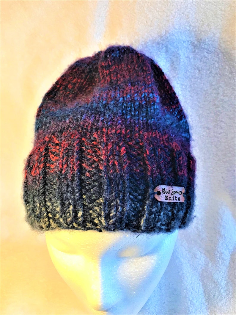May include: A hand-knitted beanie hat in a mix of colors including red, blue, and black. The hat is displayed on a white mannequin head. A small wooden tag with the text "Blue Spruce Knits" is attached to the hat.