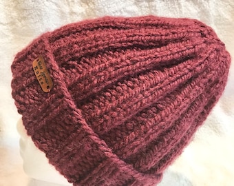 May include: A burgundy knit beanie with a ribbed design. The hat features a folded brim and a small brown tag with the text "Blue Spruce Knits". The hat is displayed on a white mannequin head.