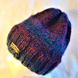 May include: A hand-knitted beanie in shades of navy, red, and blue. The hat features a tag that reads "Blue Spruce Knits". The beanie is displayed on a white mannequin head.
