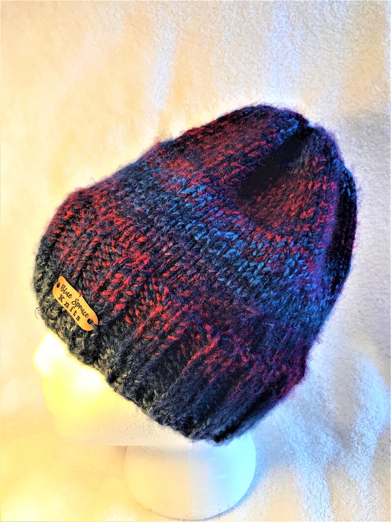 May include: A hand-knitted beanie in shades of navy, red, and blue. The hat features a tag that reads "Blue Spruce Knits". The beanie is displayed on a white mannequin head.