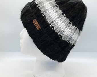 May include: A black and gray knit beanie with a diagonal stripe pattern. The hat is displayed on a white mannequin head. A small brown leather tag with the text "Blue Spruce Knits" is attached to the side.