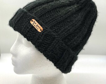 May include: A black knit beanie with a folded brim. A small, brown leather tag with the text "Blue Spruce Knits" is attached to the front of the hat. The hat is displayed on a white mannequin head.