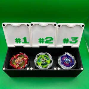 May include: A black storage box with three compartments, each labeled with a number in green. The box contains three colorful spinning tops: red and gold, green and silver, and purple and blue. The background is a solid green.