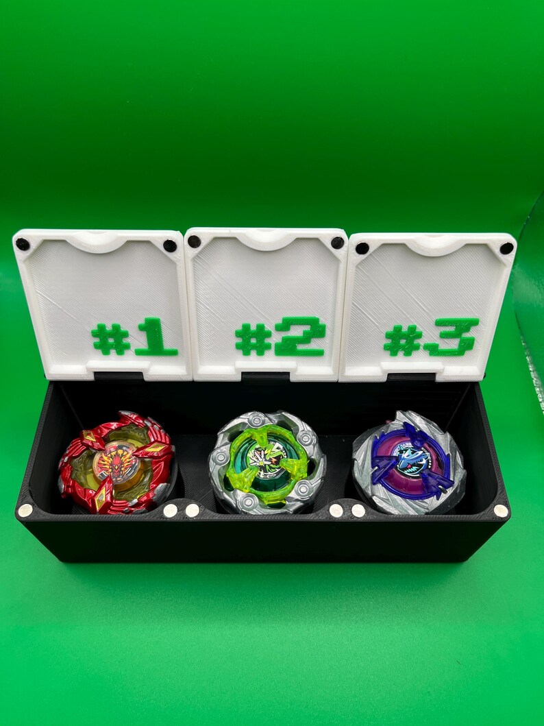 May include: A black storage box with three compartments, each labeled with a number in green. The box contains three colorful spinning tops: red and gold, green and silver, and purple and blue. The background is a solid green.