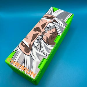 May include: A rectangular, lime green pencil case with a detailed illustration of a character with white hair and a determined expression. The case has the word "CHRIMUN" printed in orange. The background is a solid blue.