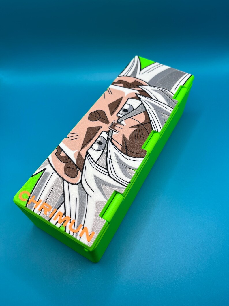 May include: A rectangular, lime green pencil case with a detailed illustration of a character with white hair and a determined expression. The case has the word "CHRIMUN" printed in orange. The background is a solid blue.