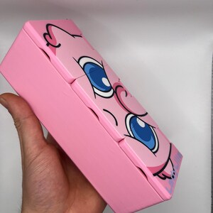 May include: A pink, rectangular box with a cartoon face design. The box features large blue eyes, pink cheeks, and a black outline. The box is held in a hand and appears to be made of wood or a similar material.