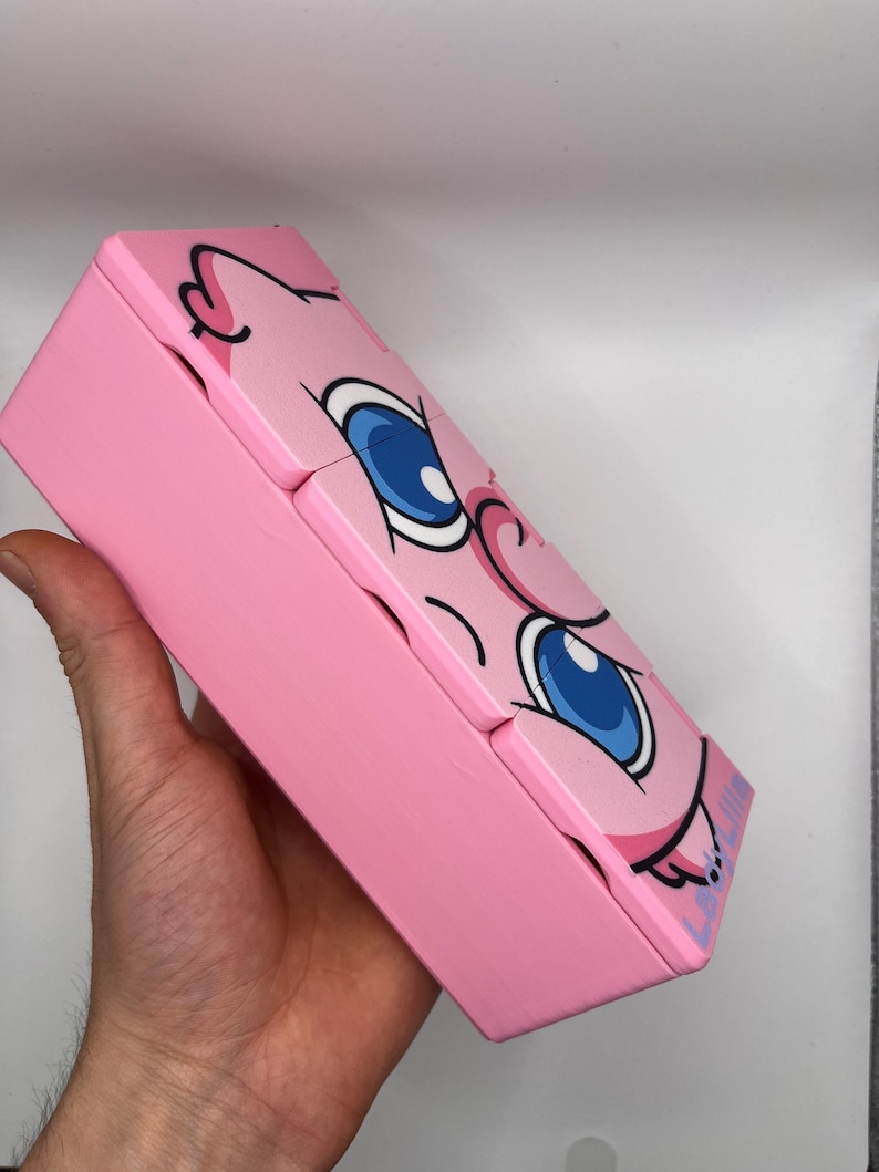 May include: A pink, rectangular box with a cartoon face design. The box features large blue eyes, pink cheeks, and a black outline. The box is held in a hand and appears to be made of wood or a similar material.