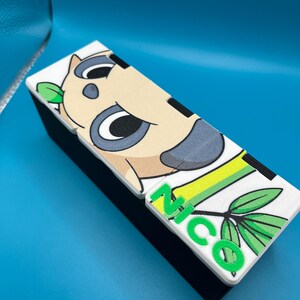 May include: A rectangular black box with a white top featuring a cartoon koala design and the name "NICO" in green letters. The koala has gray ears and a light brown face. Green leaves are also present.