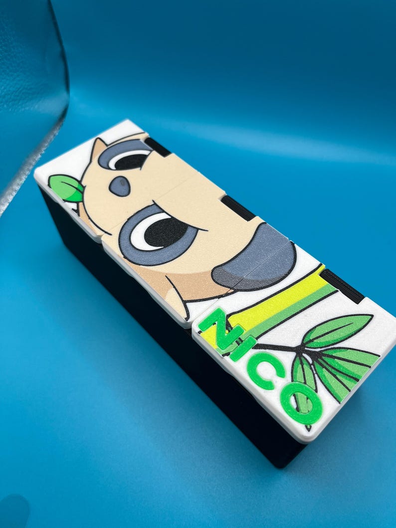 May include: A rectangular black box with a white top featuring a cartoon koala design and the name "NICO" in green letters. The koala has gray ears and a light brown face. Green leaves are also present.