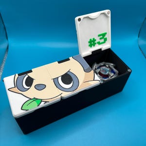 May include: A black storage box with a hinged lid, featuring a cartoon character design on the lid. The lid is open, revealing a silver and blue toy inside. The lid also has the number "#3" in green.