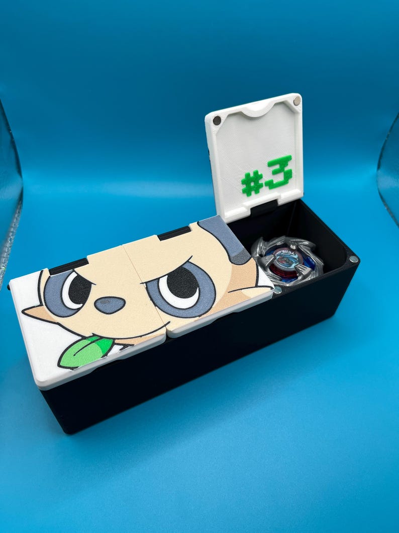 May include: A black storage box with a hinged lid, featuring a cartoon character design on the lid. The lid is open, revealing a silver and blue toy inside. The lid also has the number "#3" in green.