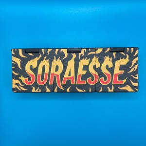 Puede incluir: Un objeto rectangular negro con la palabra "SORAESSE" en amarillo y rojo, rodeado de dise&ntilde;os en forma de llama naranja. El objeto est&aacute; sobre un fondo azul.