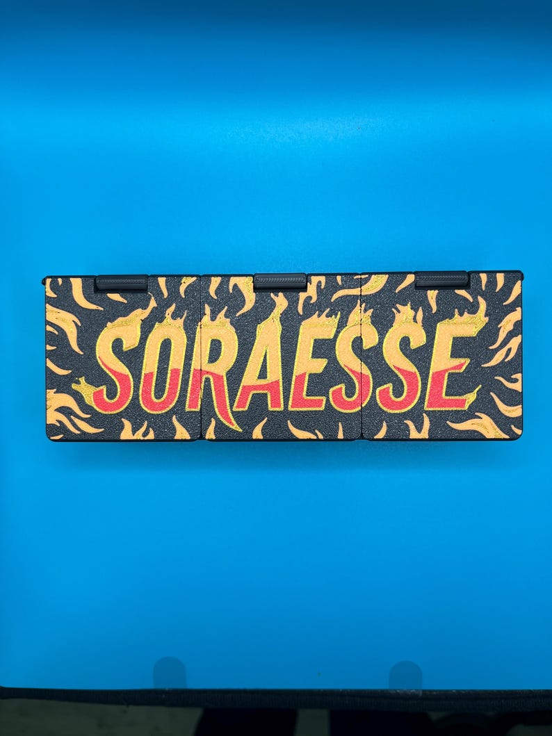 Puede incluir: Un objeto rectangular negro con la palabra "SORAESSE" en amarillo y rojo, rodeado de dise&ntilde;os en forma de llama naranja. El objeto est&aacute; sobre un fondo azul.