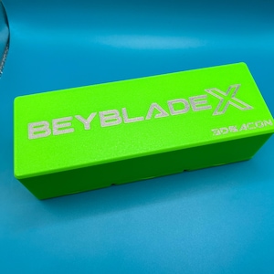 May include: A vivid green rectangular box featuring the silver text "BEYBLADE X" and "3DEACON". The box is presented against a blue backdrop, creating a striking contrast.