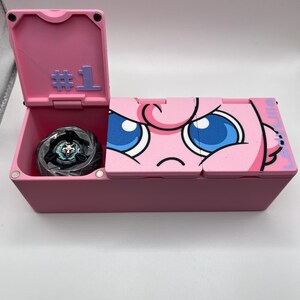 May include: A pink storage box with a hinged lid, featuring a cartoon character design on the front. The lid is open, revealing a black and silver toy inside. The box has the number "#1" on the lid.