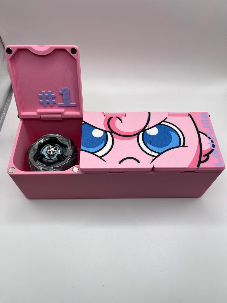 May include: A pink storage box with a hinged lid, featuring a cartoon character design on the front. The lid is open, revealing a black and silver toy inside. The box has the number "#1" on the lid.