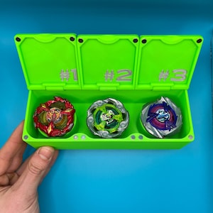May include: A vibrant green storage case divided into three sections, each marked with a number. The case contains three spinning tops in red, green, and blue. The background is a solid blue.