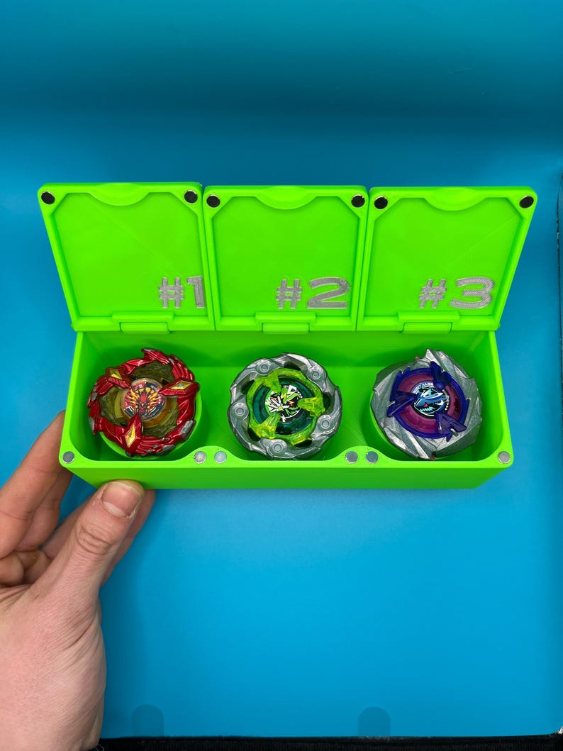 May include: A vibrant green storage case divided into three sections, each marked with a number. The case contains three spinning tops in red, green, and blue. The background is a solid blue.