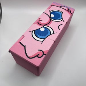 May include: A pink rectangular box with a cartoon face design. The face has large blue eyes, a pink nose, and a smiling mouth. The box is made of a solid material and has a hinged lid.