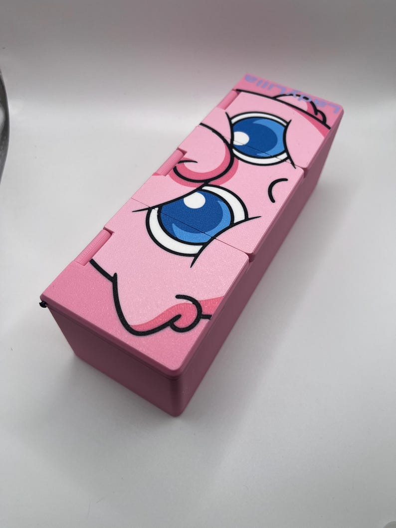 May include: A pink rectangular box with a cartoon face design. The face has large blue eyes, a pink nose, and a smiling mouth. The box is made of a solid material and has a hinged lid.