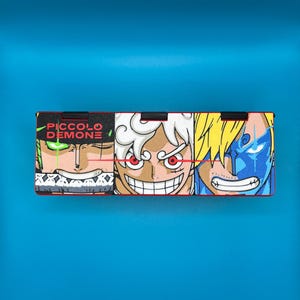 Peut inclure: Bo&icirc;te rectangulaire orn&eacute;e de trois visages de personnages d'anime aux couleurs vives et aux expressions d&eacute;taill&eacute;es. Le texte "PICCOLO DEMONE" est en vert. Le fond est bleu uni.