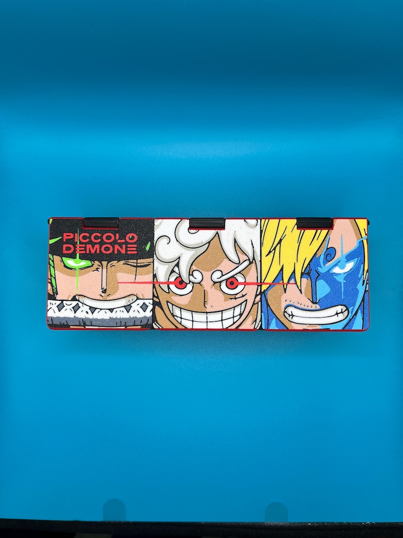 Peut inclure: Bo&icirc;te rectangulaire orn&eacute;e de trois visages de personnages d'anime aux couleurs vives et aux expressions d&eacute;taill&eacute;es. Le texte "PICCOLO DEMONE" est en vert. Le fond est bleu uni.