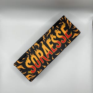 Puede incluir: Estuche rectangular negro con la palabra "SORAESSE" en llamas rojas y amarillas. El estuche tiene una bisagra negra y est&aacute; sobre un fondo blanco. Las llamas son naranjas y amarillas.