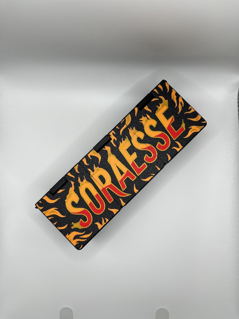 Puede incluir: Estuche rectangular negro con la palabra "SORAESSE" en llamas rojas y amarillas. El estuche tiene una bisagra negra y est&aacute; sobre un fondo blanco. Las llamas son naranjas y amarillas.