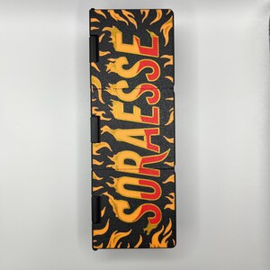 Puede incluir: Estuche rectangular negro con la palabra "SORAEESE" en letras amarillas y rojas con forma de llama. El estuche tiene una superficie texturizada y est&aacute; decorado con gr&aacute;ficos de llamas amarillas y naranjas.