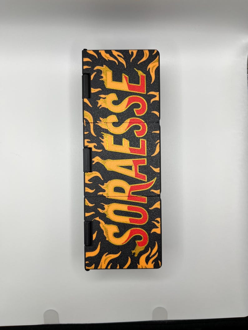 Puede incluir: Estuche rectangular negro con la palabra "SORAEESE" en letras amarillas y rojas con forma de llama. El estuche tiene una superficie texturizada y est&aacute; decorado con gr&aacute;ficos de llamas amarillas y naranjas.