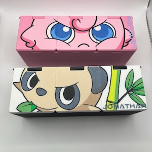 May include: Two decorative boxes, one pink with a cartoon face and blue eyes, the other black and white with a panda design and the name "JONATHAN". The boxes are likely for storage or display.