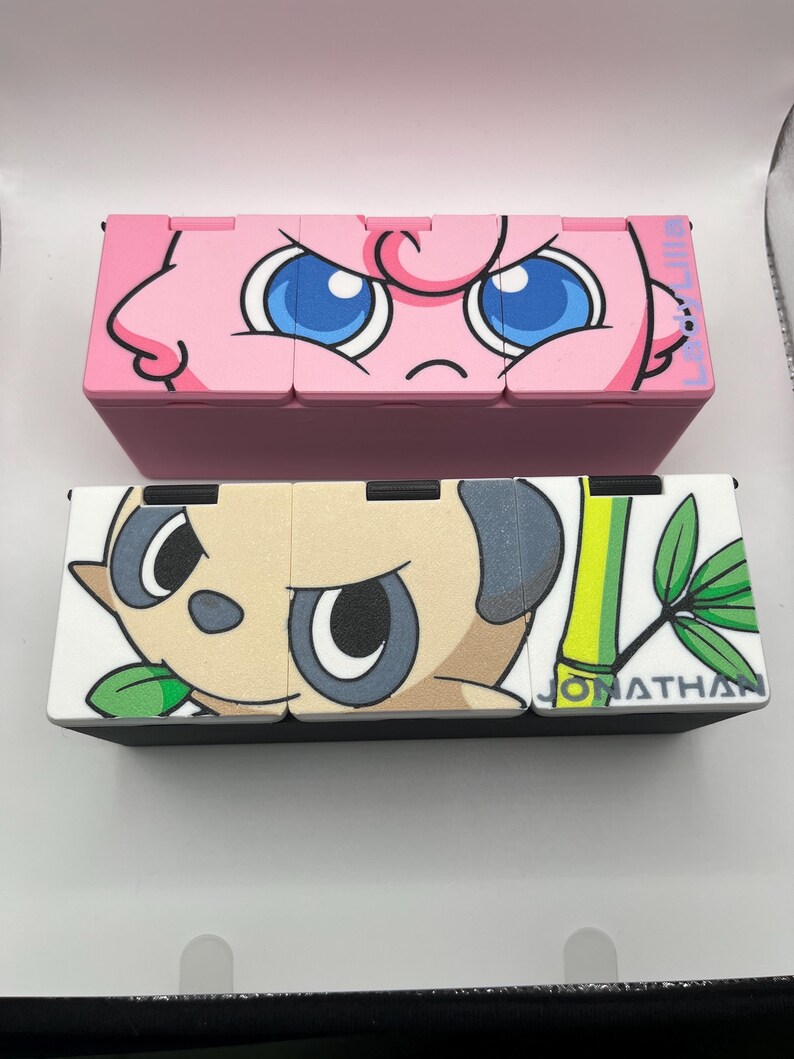 May include: Two decorative boxes, one pink with a cartoon face and blue eyes, the other black and white with a panda design and the name "JONATHAN". The boxes are likely for storage or display.
