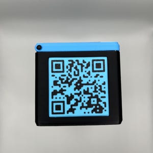 Może przedstawiać: Czarne kwadratowe urządzenie z niebieskim kodem QR na przodzie. G&oacute;rna część urządzenia jest niebieska, a kod QR ma jaśniejszy odcień niebieskiego. Urządzenie jest prawdopodobnie używane do skanowania lub wyświetlania informacji.