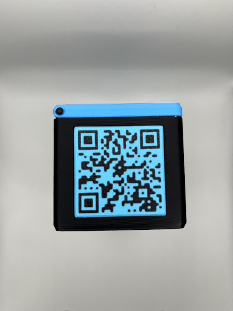 Może przedstawiać: Czarne kwadratowe urządzenie z niebieskim kodem QR na przodzie. G&oacute;rna część urządzenia jest niebieska, a kod QR ma jaśniejszy odcień niebieskiego. Urządzenie jest prawdopodobnie używane do skanowania lub wyświetlania informacji.